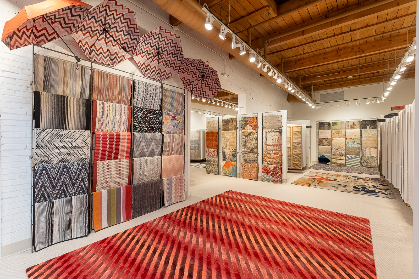 Display of various patterned rugs in a showroom with wooden ceiling and white walls.